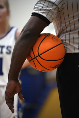 A basketball referee holds an orange basketball under their arm. They are wearing a black and white striped shirt, indicative of a referee's uniform. A player in a white jersey with blue trim is visible in the background, slightly out of focus.