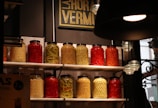 A collection of large jars filled with a variety of preserved vegetables including red peppers, yellow beans, and carrots, arranged on white shelves against a tiled wall. A sign with the words 'La Hora Vermut' is displayed above the jars. Ambient lighting creates a cozy atmosphere, with a black lampshade visible in the foreground.