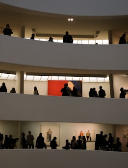 Several floors of an art museum with visitors walking and viewing artwork. The architecture features curved white walls and layered balconies. Silhouettes of people are visible against brightly lit gallery spaces displaying paintings, including a prominent red painting.