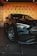 A sleek black car with shiny chrome accents is parked indoors. The car is positioned in front of a wall with repeating 'GA' branding on a warm-toned backdrop. The floor is tiled with large, dark tiles, enhancing the modern vibe of the scene.