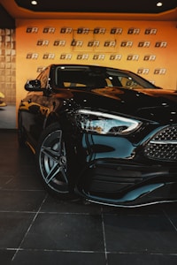 A sleek black car with shiny chrome accents is parked indoors. The car is positioned in front of a wall with repeating 'GA' branding on a warm-toned backdrop. The floor is tiled with large, dark tiles, enhancing the modern vibe of the scene.