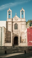 A historic stone facade of a church with a large, arched entrance, flanked by two bell towers. The architecture features detailed carvings and wrought iron fencing. The facade is adorned with a stained glass window and the words 'Ave Maria' inscribed in a circular recess. The sky is clear blue and the building is partially shaded.