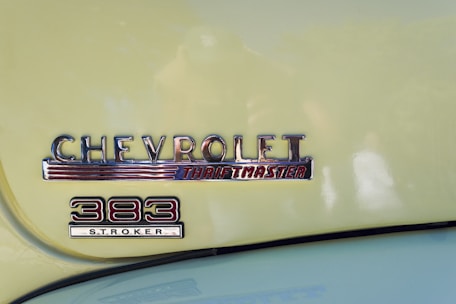 A classic car badge with the embossed text 'CHEVROLET Thriftmaster' and '383 Stroker' displayed on a light yellow surface. The badge features a retro style design with chrome finish and red accents.