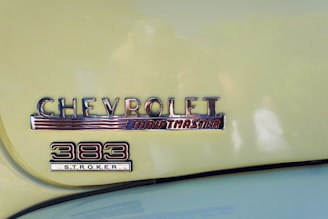 A classic car badge with the embossed text 'CHEVROLET Thriftmaster' and '383 Stroker' displayed on a light yellow surface. The badge features a retro style design with chrome finish and red accents.