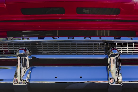 The front grille of a vintage Chevrolet vehicle features prominently, with streamlined chrome details and a bold red backdrop. The chrome is shiny and reflective, adding a classic and polished look.