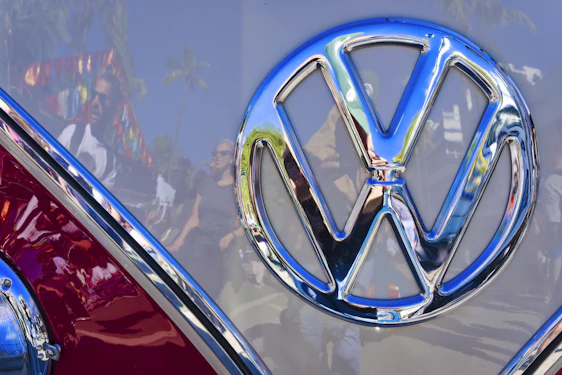 A close-up of a shiny Volkswagen logo is prominently featured in the center. The chrome emblem reflects the surroundings, including people, palm trees, and vibrant flags, indicating an outdoor event or fair.