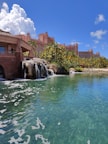A tropical resort scene featuring a large, pink-colored hotel with spires, surrounded by palm trees and lush greenery. A serene, crystal-clear turquoise pond is in the foreground, accented by small waterfalls cascading from rocks. The sky is bright blue with large, fluffy white clouds.