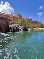 A tropical resort scene featuring a large, pink-colored hotel with spires, surrounded by palm trees and lush greenery. A serene, crystal-clear turquoise pond is in the foreground, accented by small waterfalls cascading from rocks. The sky is bright blue with large, fluffy white clouds.