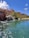 A tropical resort scene featuring a large, pink-colored hotel with spires, surrounded by palm trees and lush greenery. A serene, crystal-clear turquoise pond is in the foreground, accented by small waterfalls cascading from rocks. The sky is bright blue with large, fluffy white clouds.