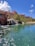A tropical resort scene featuring a large, pink-colored hotel with spires, surrounded by palm trees and lush greenery. A serene, crystal-clear turquoise pond is in the foreground, accented by small waterfalls cascading from rocks. The sky is bright blue with large, fluffy white clouds.