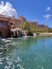 A tropical resort scene featuring a large, pink-colored hotel with spires, surrounded by palm trees and lush greenery. A serene, crystal-clear turquoise pond is in the foreground, accented by small waterfalls cascading from rocks. The sky is bright blue with large, fluffy white clouds.