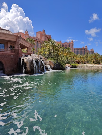 A tropical resort scene featuring a large, pink-colored hotel with spires, surrounded by palm trees and lush greenery. A serene, crystal-clear turquoise pond is in the foreground, accented by small waterfalls cascading from rocks. The sky is bright blue with large, fluffy white clouds.