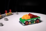 A small toy race car, predominantly green with vibrant red wheels, is positioned on a white surface. The body of the car is decorated with multicolored blocks, including yellow and red pieces on top. In the background, there are several gray toy blocks and a barricade with red lights.