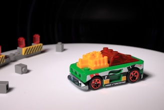 A small toy race car, predominantly green with vibrant red wheels, is positioned on a white surface. The body of the car is decorated with multicolored blocks, including yellow and red pieces on top. In the background, there are several gray toy blocks and a barricade with red lights.