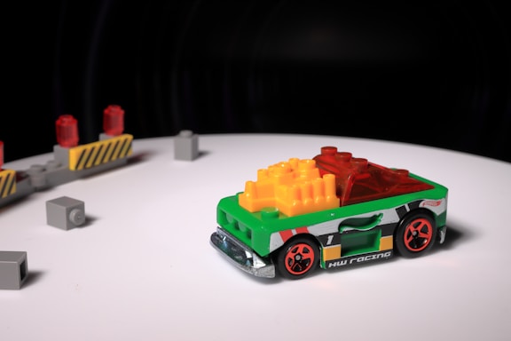 A small toy race car, predominantly green with vibrant red wheels, is positioned on a white surface. The body of the car is decorated with multicolored blocks, including yellow and red pieces on top. In the background, there are several gray toy blocks and a barricade with red lights.