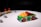 A small toy race car, predominantly green with vibrant red wheels, is positioned on a white surface. The body of the car is decorated with multicolored blocks, including yellow and red pieces on top. In the background, there are several gray toy blocks and a barricade with red lights.