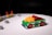A small toy race car, predominantly green with vibrant red wheels, is positioned on a white surface. The body of the car is decorated with multicolored blocks, including yellow and red pieces on top. In the background, there are several gray toy blocks and a barricade with red lights.