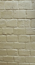 A textured brick wall with a pale, beige-colored painted surface. The bricks appear rough and slightly worn, with uneven application of paint, highlighting the individual texture of each block. Horizontal and vertical lines separate the bricks, giving a structured pattern to the entire wall.