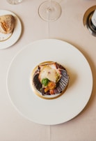 A gourmet dish is artfully arranged on a white plate with a scallop shell containing scallops, a seared garnish, and garnishes. This is set on a beige tablecloth, with a wine glass, a piece of bread on a small plate, and a portion of a wine bottle also visible.