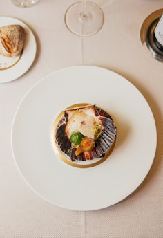 A gourmet dish is artfully arranged on a white plate with a scallop shell containing scallops, a seared garnish, and garnishes. This is set on a beige tablecloth, with a wine glass, a piece of bread on a small plate, and a portion of a wine bottle also visible.