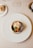 A gourmet dish is artfully arranged on a white plate with a scallop shell containing scallops, a seared garnish, and garnishes. This is set on a beige tablecloth, with a wine glass, a piece of bread on a small plate, and a portion of a wine bottle also visible.