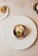 A gourmet dish is artfully arranged on a white plate with a scallop shell containing scallops, a seared garnish, and garnishes. This is set on a beige tablecloth, with a wine glass, a piece of bread on a small plate, and a portion of a wine bottle also visible.