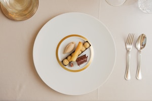 A gourmet dessert arrangement is plated on a large white plate with a gold rim. The dessert features a variety of textures and elements, including a scoop of caramel-colored ice cream, a rolled biscuit, chocolate garnishes, and small cream swirls. The setting includes polished silver cutlery on a neutral tablecloth, and a clear glass positioned at the top left.