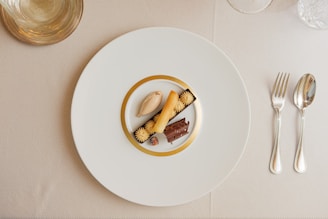 A gourmet dessert arrangement is plated on a large white plate with a gold rim. The dessert features a variety of textures and elements, including a scoop of caramel-colored ice cream, a rolled biscuit, chocolate garnishes, and small cream swirls. The setting includes polished silver cutlery on a neutral tablecloth, and a clear glass positioned at the top left.