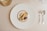 A gourmet dessert arrangement is plated on a large white plate with a gold rim. The dessert features a variety of textures and elements, including a scoop of caramel-colored ice cream, a rolled biscuit, chocolate garnishes, and small cream swirls. The setting includes polished silver cutlery on a neutral tablecloth, and a clear glass positioned at the top left.