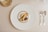 A gourmet dessert arrangement is plated on a large white plate with a gold rim. The dessert features a variety of textures and elements, including a scoop of caramel-colored ice cream, a rolled biscuit, chocolate garnishes, and small cream swirls. The setting includes polished silver cutlery on a neutral tablecloth, and a clear glass positioned at the top left.