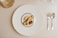 A gourmet dessert arrangement is plated on a large white plate with a gold rim. The dessert features a variety of textures and elements, including a scoop of caramel-colored ice cream, a rolled biscuit, chocolate garnishes, and small cream swirls. The setting includes polished silver cutlery on a neutral tablecloth, and a clear glass positioned at the top left.