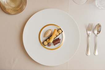 A gourmet dessert arrangement is plated on a large white plate with a gold rim. The dessert features a variety of textures and elements, including a scoop of caramel-colored ice cream, a rolled biscuit, chocolate garnishes, and small cream swirls. The setting includes polished silver cutlery on a neutral tablecloth, and a clear glass positioned at the top left.
