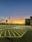 A parking lot at sunset, with the sky transitioning from deep blue at the top to a vibrant orange near the horizon. Trees without leaves are silhouetted against the sky, and several cars are parked in the lot. The area features clear yellow-lined parking spaces and a tall light pole.