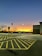 A parking lot at sunset, with the sky transitioning from deep blue at the top to a vibrant orange near the horizon. Trees without leaves are silhouetted against the sky, and several cars are parked in the lot. The area features clear yellow-lined parking spaces and a tall light pole.