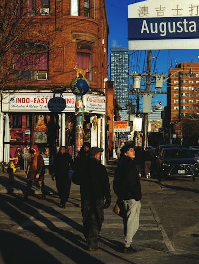 Urban street scene at an intersection, featuring a brick building with a store called Indo-East Trading. Pedestrians are walking in different directions under a clear blue sky. A street sign displaying 'Augusta' is prominently visible along with power lines and high-rise buildings in the background.