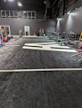 A spacious gym with a black and white color scheme featuring various fitness equipment. On the left, numerous weight plates and bars are organized on racks. In the center, a large open workout area is visible with a textured floor and white directional markings. To the right, there are treadmills, stationary bikes, and other workout machines along with large exercise balls. Exercise bands and weightlifting belts are hanging on hooks at the back wall. A single person is seen near a door, adding a human element to the scene.
