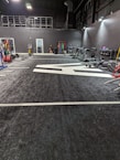 A spacious gym with a black and white color scheme featuring various fitness equipment. On the left, numerous weight plates and bars are organized on racks. In the center, a large open workout area is visible with a textured floor and white directional markings. To the right, there are treadmills, stationary bikes, and other workout machines along with large exercise balls. Exercise bands and weightlifting belts are hanging on hooks at the back wall. A single person is seen near a door, adding a human element to the scene.