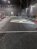 A spacious gym with a black and white color scheme featuring various fitness equipment. On the left, numerous weight plates and bars are organized on racks. In the center, a large open workout area is visible with a textured floor and white directional markings. To the right, there are treadmills, stationary bikes, and other workout machines along with large exercise balls. Exercise bands and weightlifting belts are hanging on hooks at the back wall. A single person is seen near a door, adding a human element to the scene.