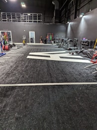 A spacious gym with a black and white color scheme featuring various fitness equipment. On the left, numerous weight plates and bars are organized on racks. In the center, a large open workout area is visible with a textured floor and white directional markings. To the right, there are treadmills, stationary bikes, and other workout machines along with large exercise balls. Exercise bands and weightlifting belts are hanging on hooks at the back wall. A single person is seen near a door, adding a human element to the scene.
