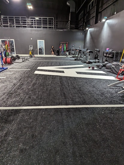 A spacious gym with a black and white color scheme featuring various fitness equipment. On the left, numerous weight plates and bars are organized on racks. In the center, a large open workout area is visible with a textured floor and white directional markings. To the right, there are treadmills, stationary bikes, and other workout machines along with large exercise balls. Exercise bands and weightlifting belts are hanging on hooks at the back wall. A single person is seen near a door, adding a human element to the scene.