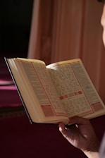 A person is holding an open book, possibly a religious text, with intricate and decorative script. The setting appears to be serene with soft lighting, creating a focused and contemplative atmosphere.