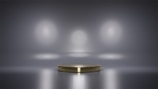 A metallic platform is centered in a minimalistic setting with a reflective surface. Soft, diffused lighting creates circular patterns on the background wall, enhancing the focus on the central stage. The ambiance is sleek and modern, with a hint of elegance.
