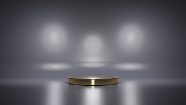 A metallic platform is centered in a minimalistic setting with a reflective surface. Soft, diffused lighting creates circular patterns on the background wall, enhancing the focus on the central stage. The ambiance is sleek and modern, with a hint of elegance.