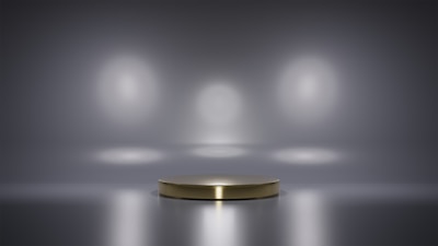 A metallic platform is centered in a minimalistic setting with a reflective surface. Soft, diffused lighting creates circular patterns on the background wall, enhancing the focus on the central stage. The ambiance is sleek and modern, with a hint of elegance.