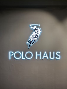 A glowing sign features an illustration of a polo player on a horse, swinging a mallet. Below the image, the words 'POLO HAUS' are illuminated in bright blue neon against a dark background.