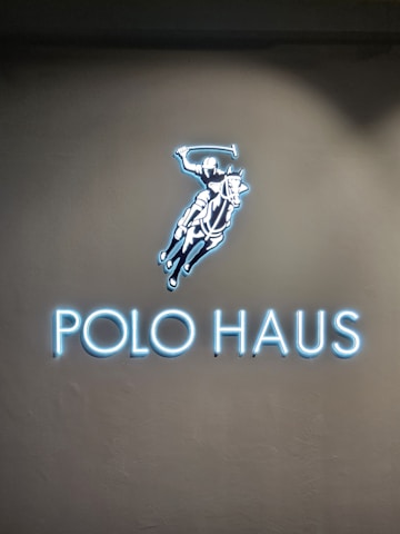 A glowing sign features an illustration of a polo player on a horse, swinging a mallet. Below the image, the words 'POLO HAUS' are illuminated in bright blue neon against a dark background.