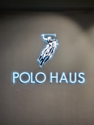 A glowing sign features an illustration of a polo player on a horse, swinging a mallet. Below the image, the words 'POLO HAUS' are illuminated in bright blue neon against a dark background.