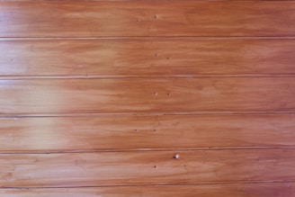 Wooden panels with a natural brown finish showing texture and subtle grain details. The surface is smooth with visible nail holes positioned in rows and slight variations in color and shading across the wood grains.