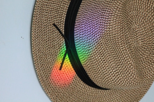 A woven straw hat is adorned with a black ribbon and displays a spectrum of colors—red, green, blue, and purple—reflecting onto its surface.