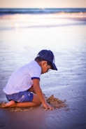 Sun-hatted toddler building a sandcastle on a sunny beach with clear blue waves in the background.